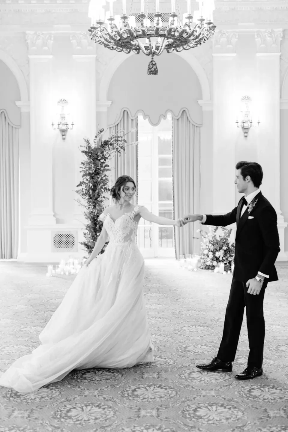 The groom takes the bride’s hand in the ballroom beneath the chandelier. Black and white.
