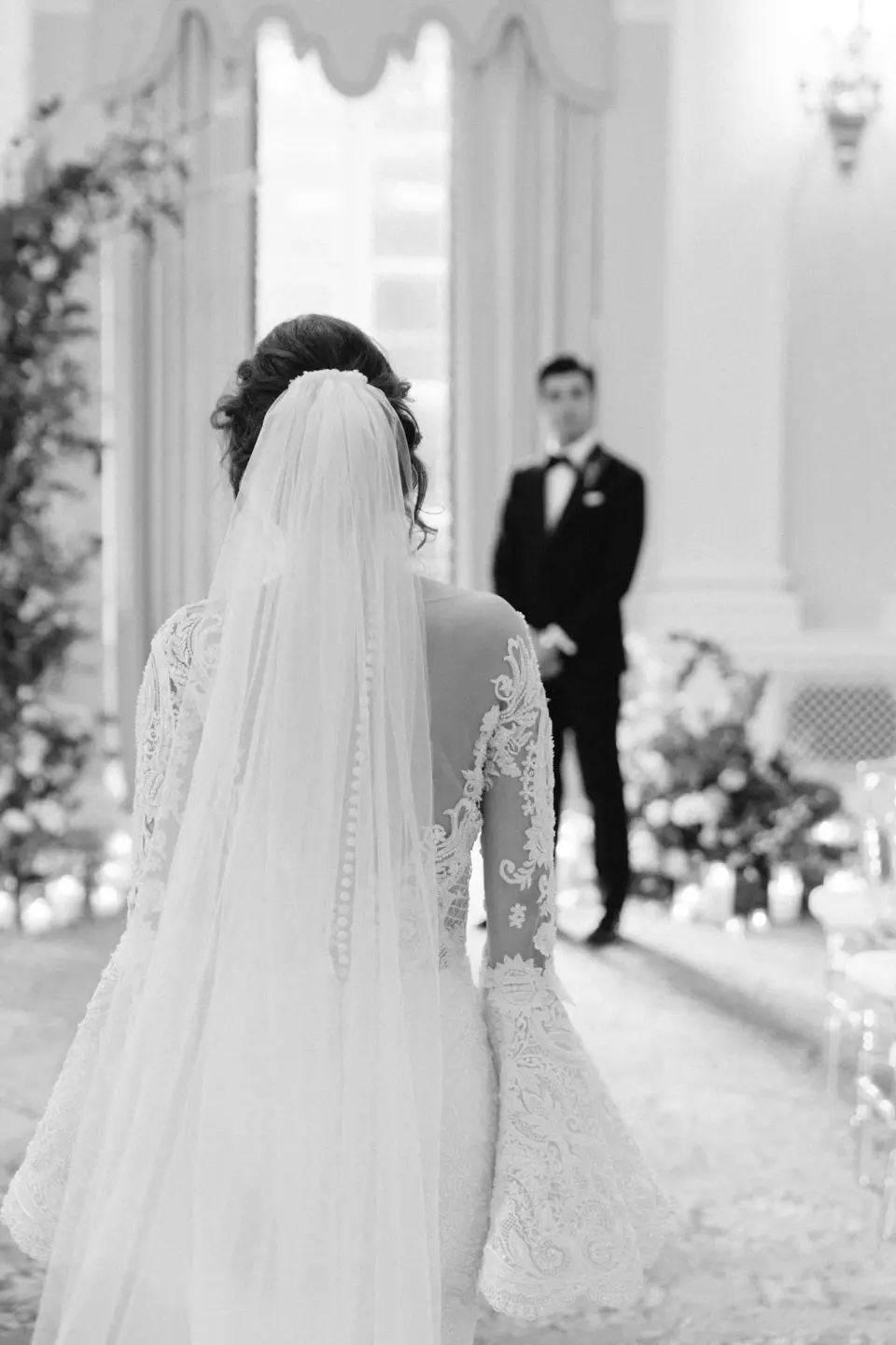 Seen from behind, the bride in a long lace veil walks toward the groom. Black and white.