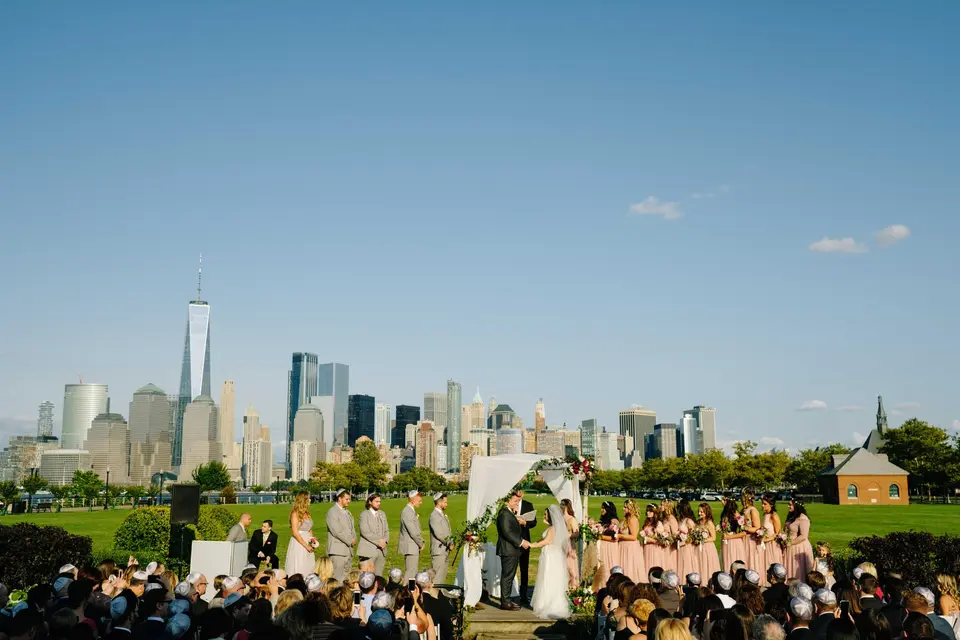 A ceremony on a lawn, guests seated before the wedding party, the Lower Manhattan skyline across the water.