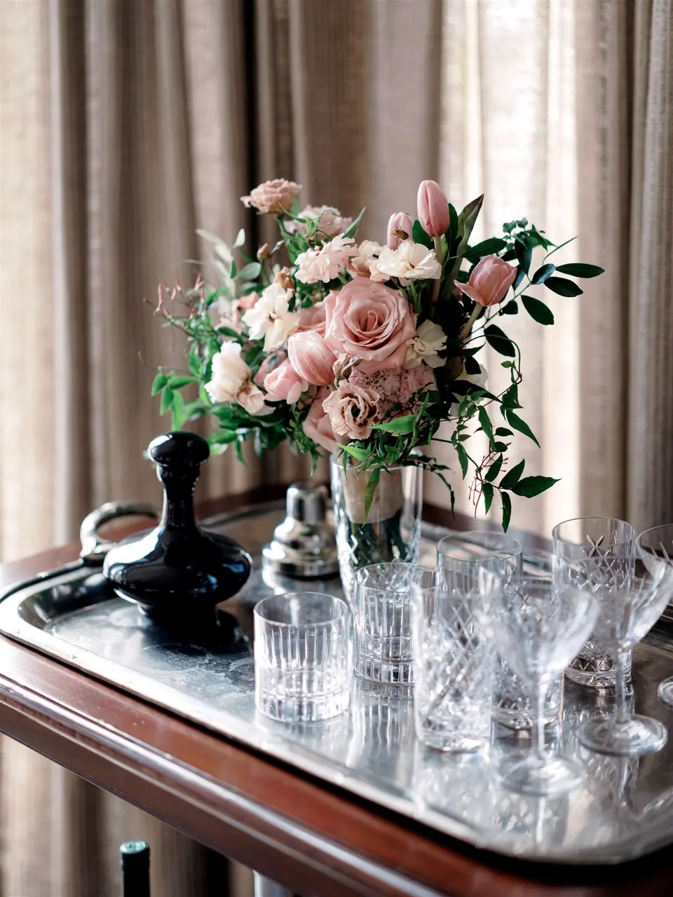 Pink roses and tulips in a glass vase on a silver bar tray with crystal glasses and a decanter.