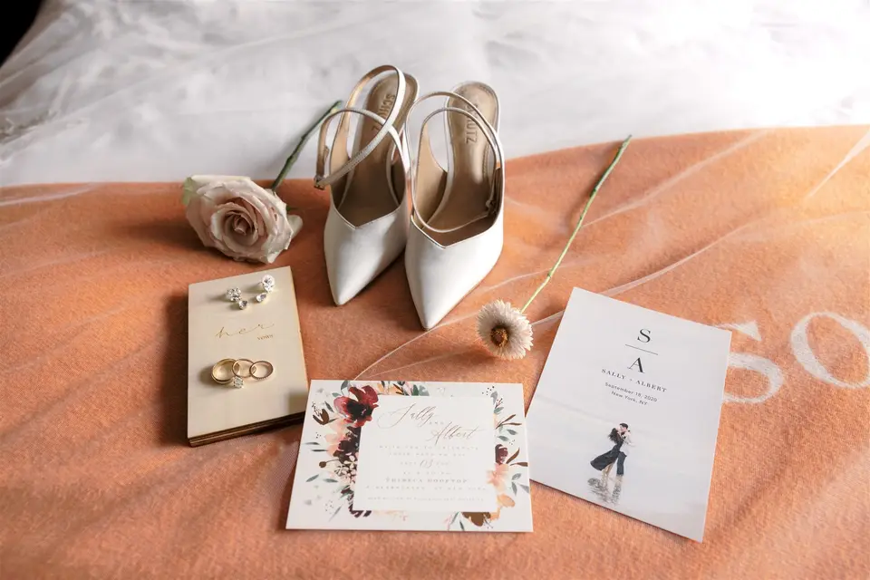White heels, the rings, a vow book and the invitation suite laid on a coral blanket.