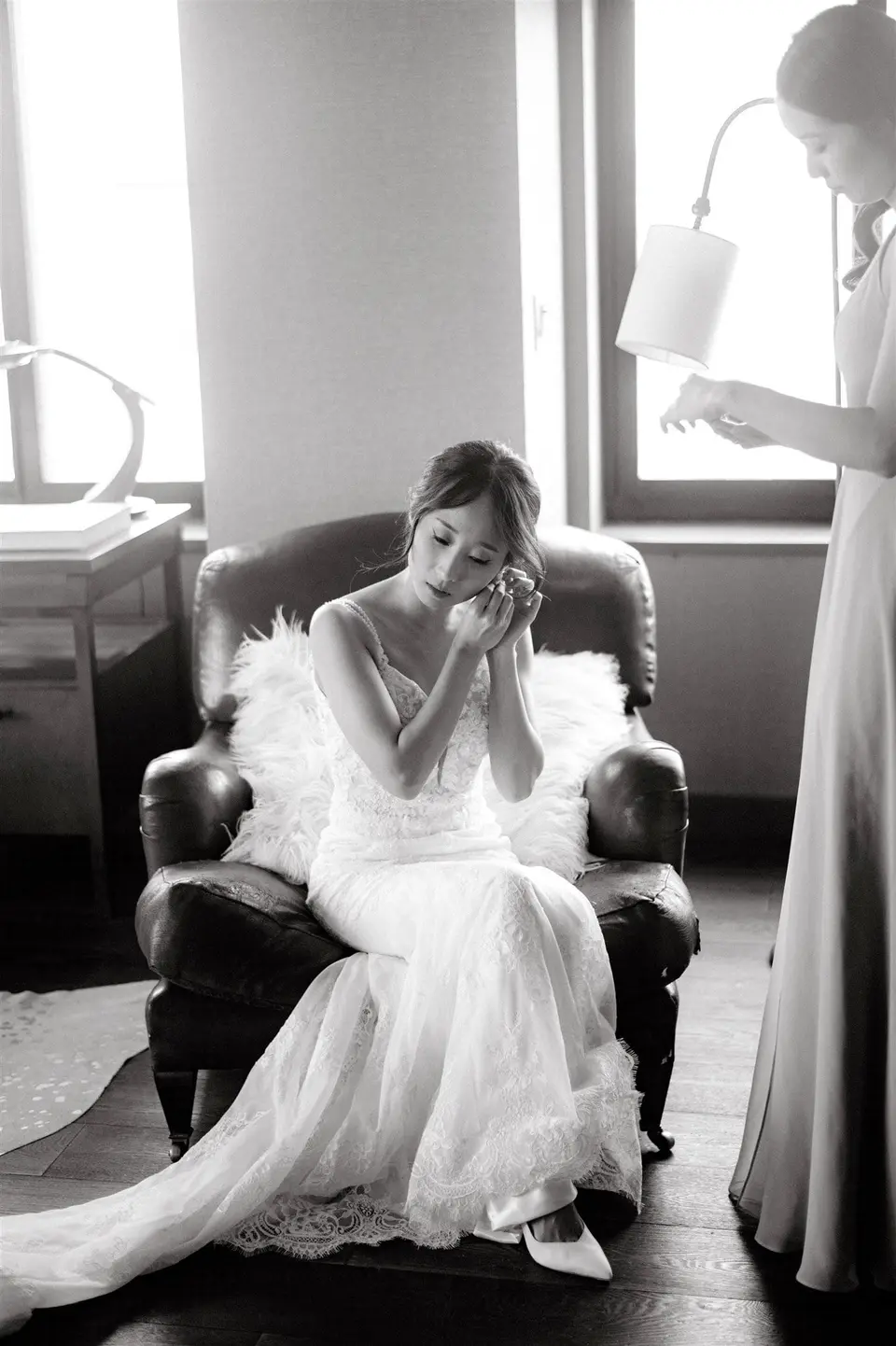 The bride puts on her earrings in a leather armchair by the window. Black and white.