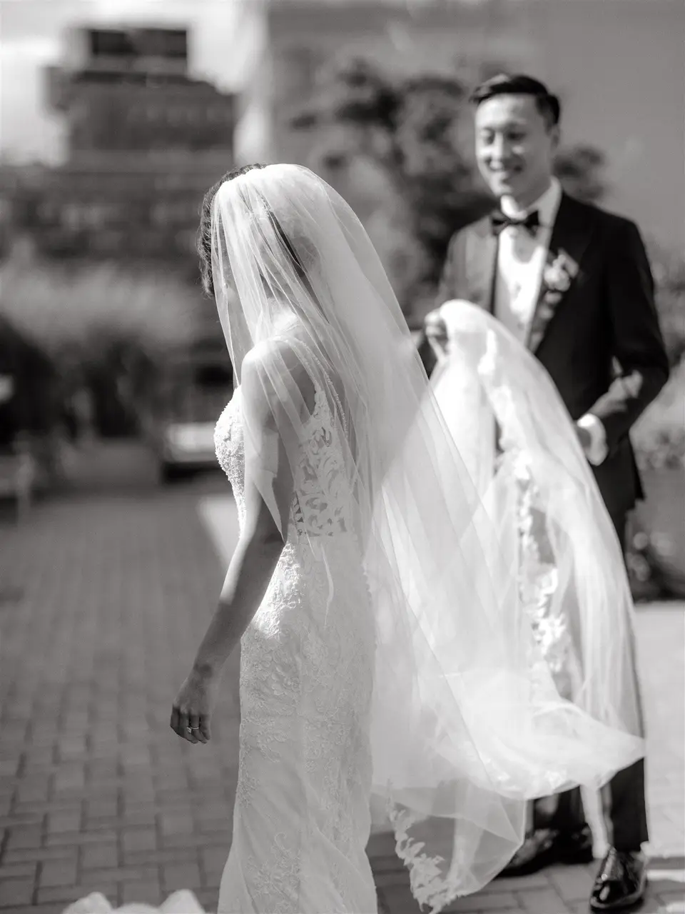 The groom smiles as the bride walks toward him, her long veil trailing. Black and white.