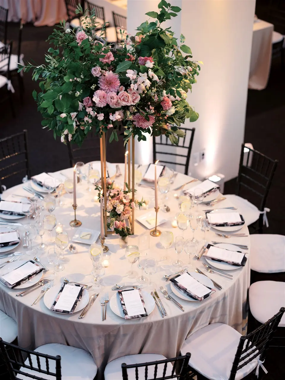 A round table beneath a tall arrangement of pink roses and greenery on a gold stand.