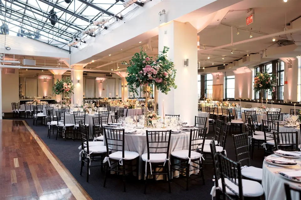 The dining room beneath the glass roof, round tables and tall flower arrangements around the dance floor.