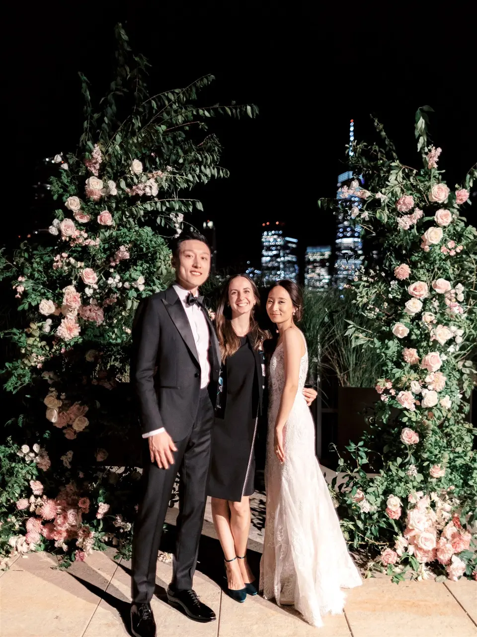 At night, the bride and groom stand with a guest between the flower arches, lit towers behind them.