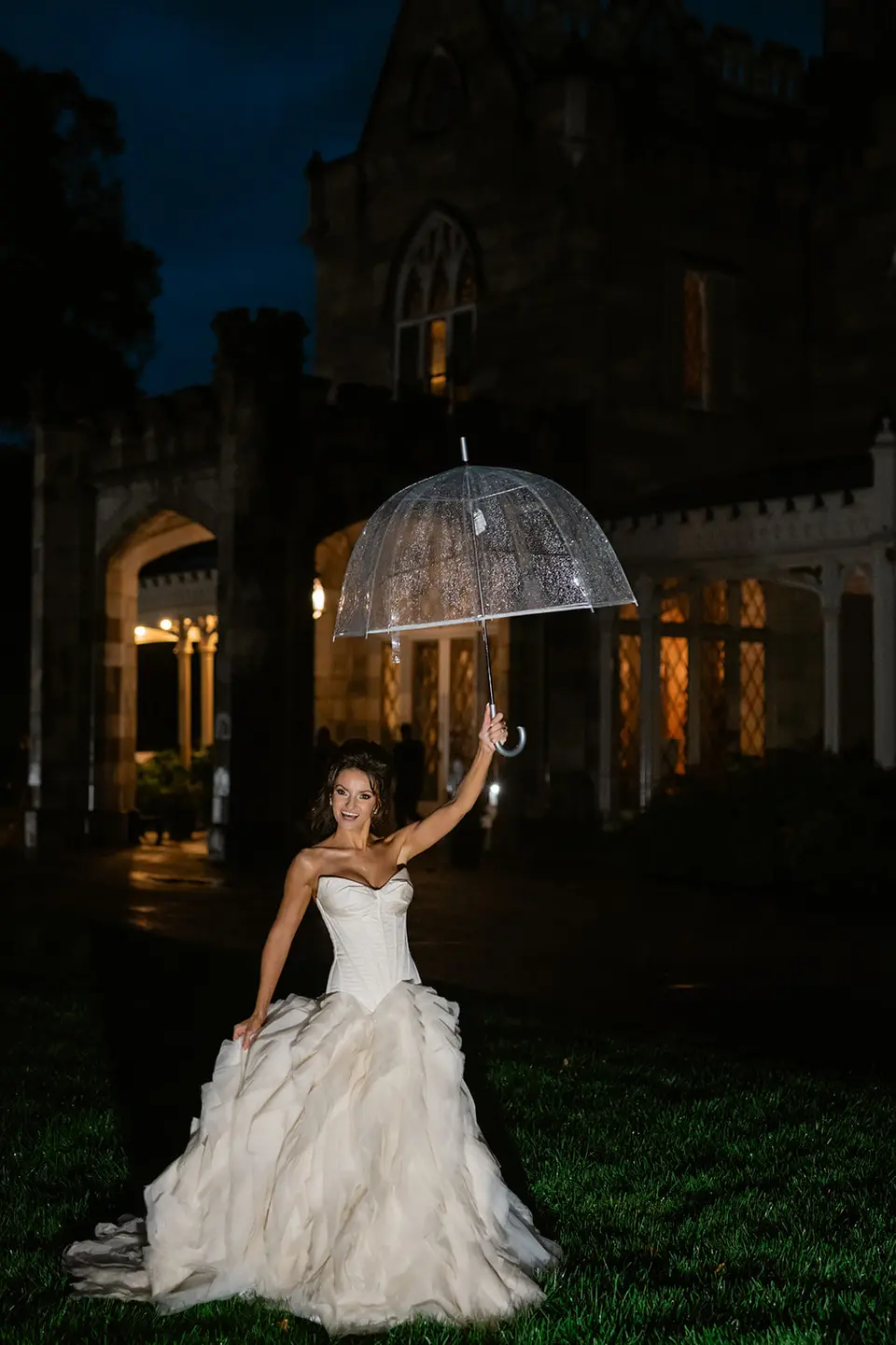 At night, the bride laughs beneath a clear umbrella on the lawn, the dark mansion behind her.
