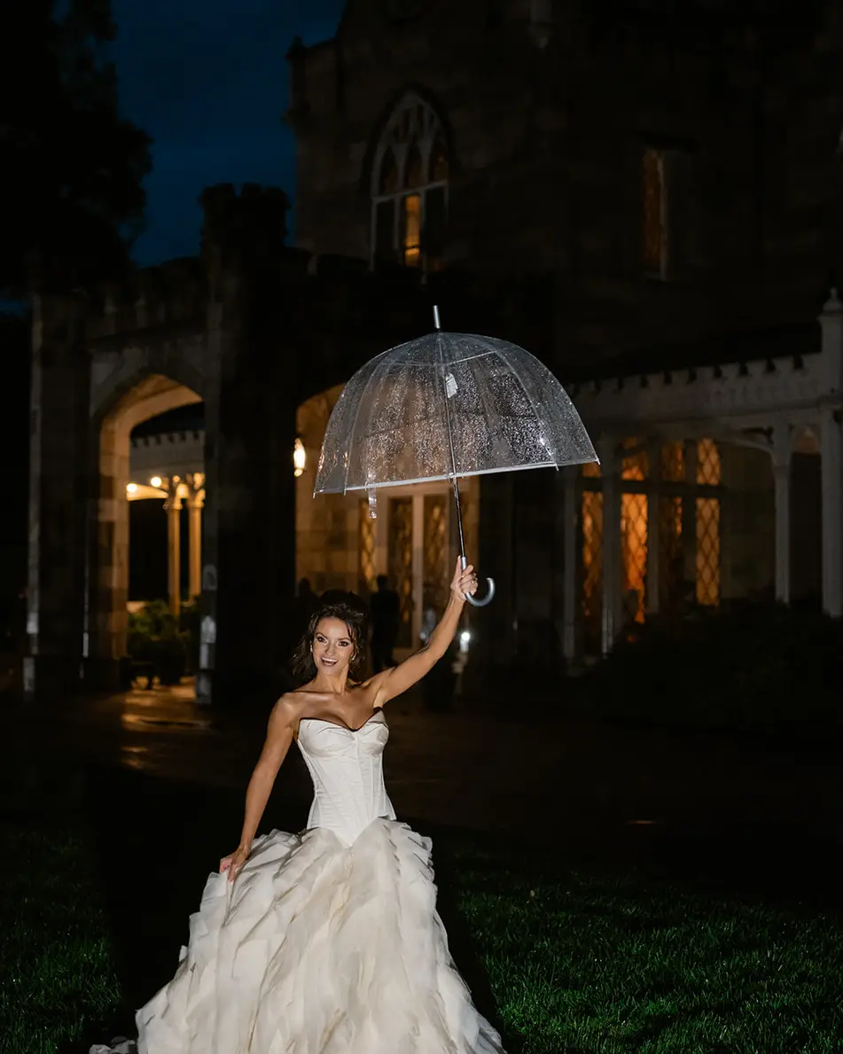 At night, the bride laughs beneath a clear umbrella on the lawn, the dark mansion behind her.