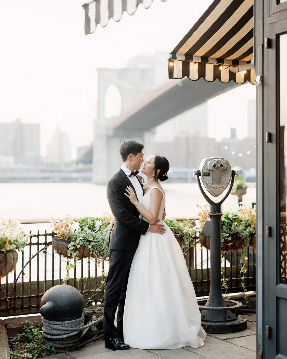 The couple kiss beneath a striped awning by the water, the Brooklyn Bridge soft in the haze behind them.