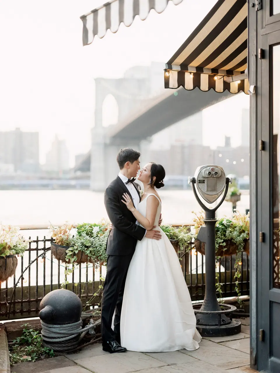 The couple kiss beneath a striped awning by the water, the Brooklyn Bridge soft in the haze behind them.