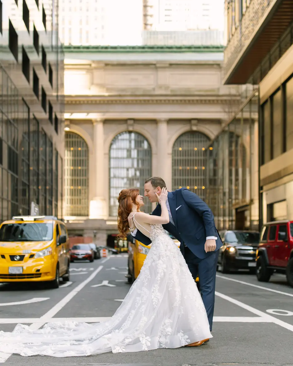 A bride and groom kiss in the middle of Vanderbilt Avenue, Grand Central Terminal behind them and a yellow cab waiting at the light.
