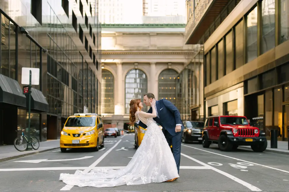 A bride and groom kiss in the middle of Vanderbilt Avenue, Grand Central Terminal behind them and a yellow cab waiting at the light.