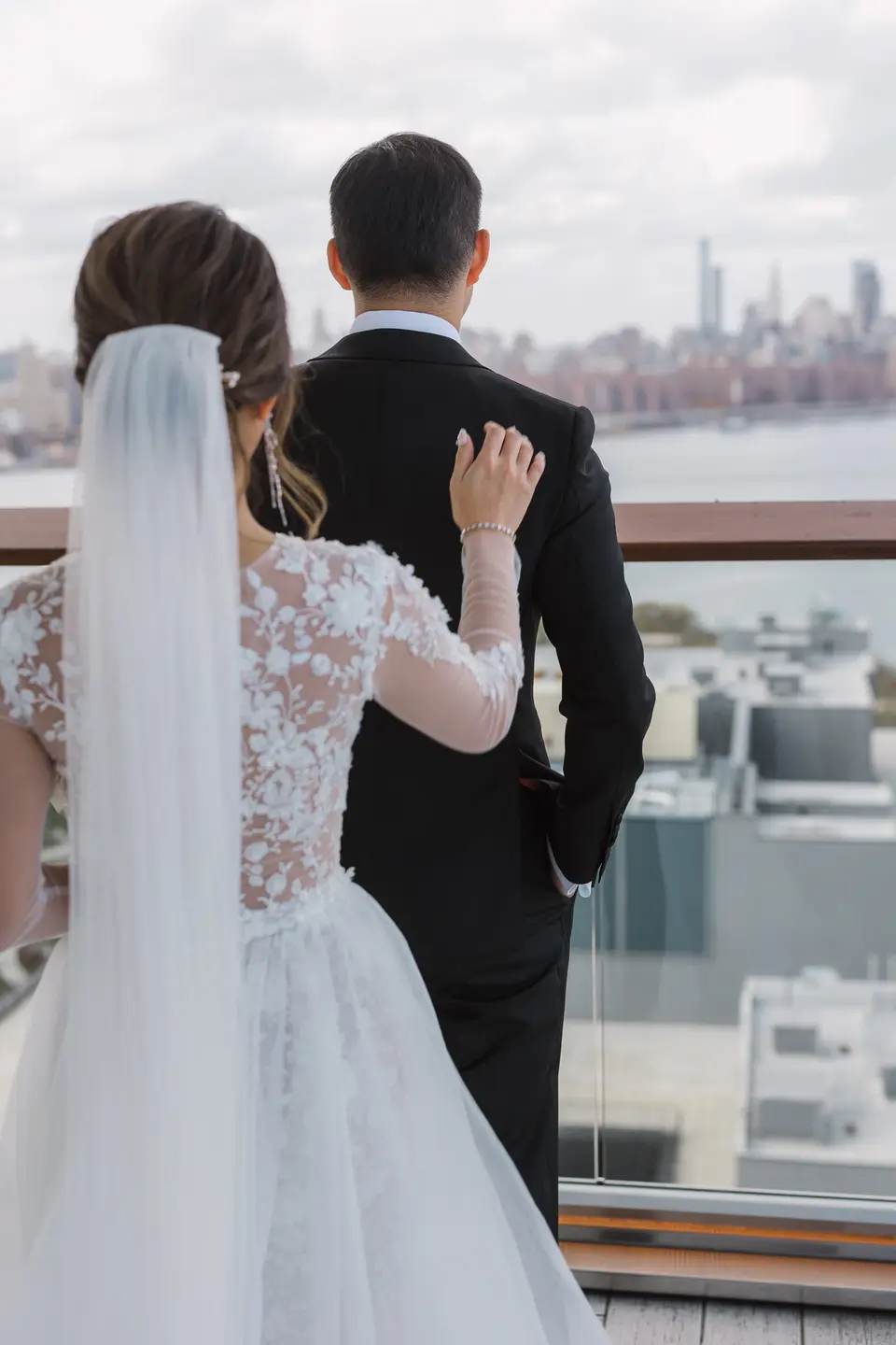 From behind, the bride rests a hand on the groom's shoulder as they look out over Brooklyn to the Manhattan skyline.