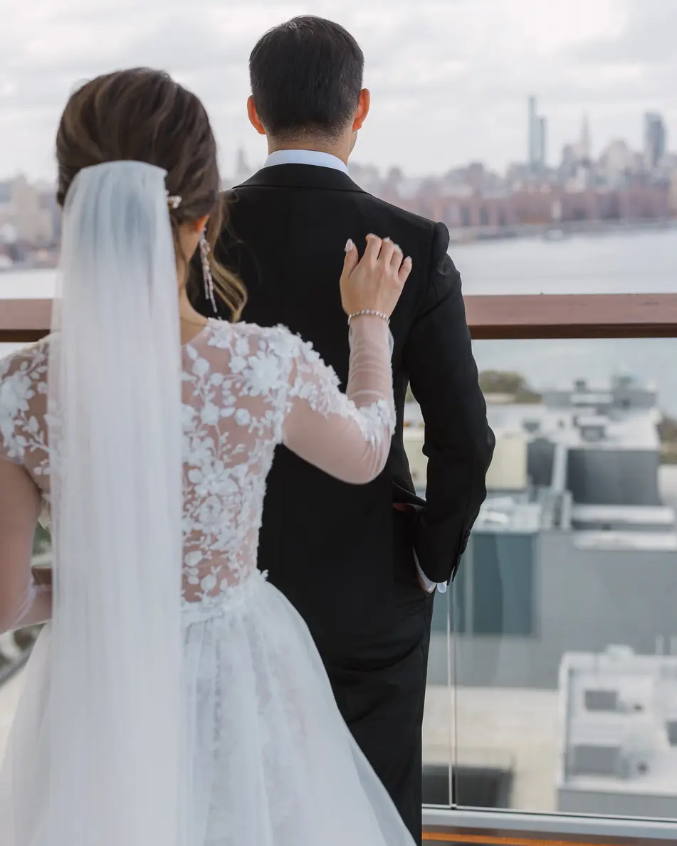 From behind, the bride rests a hand on the groom's shoulder as they look out over Brooklyn to the Manhattan skyline.