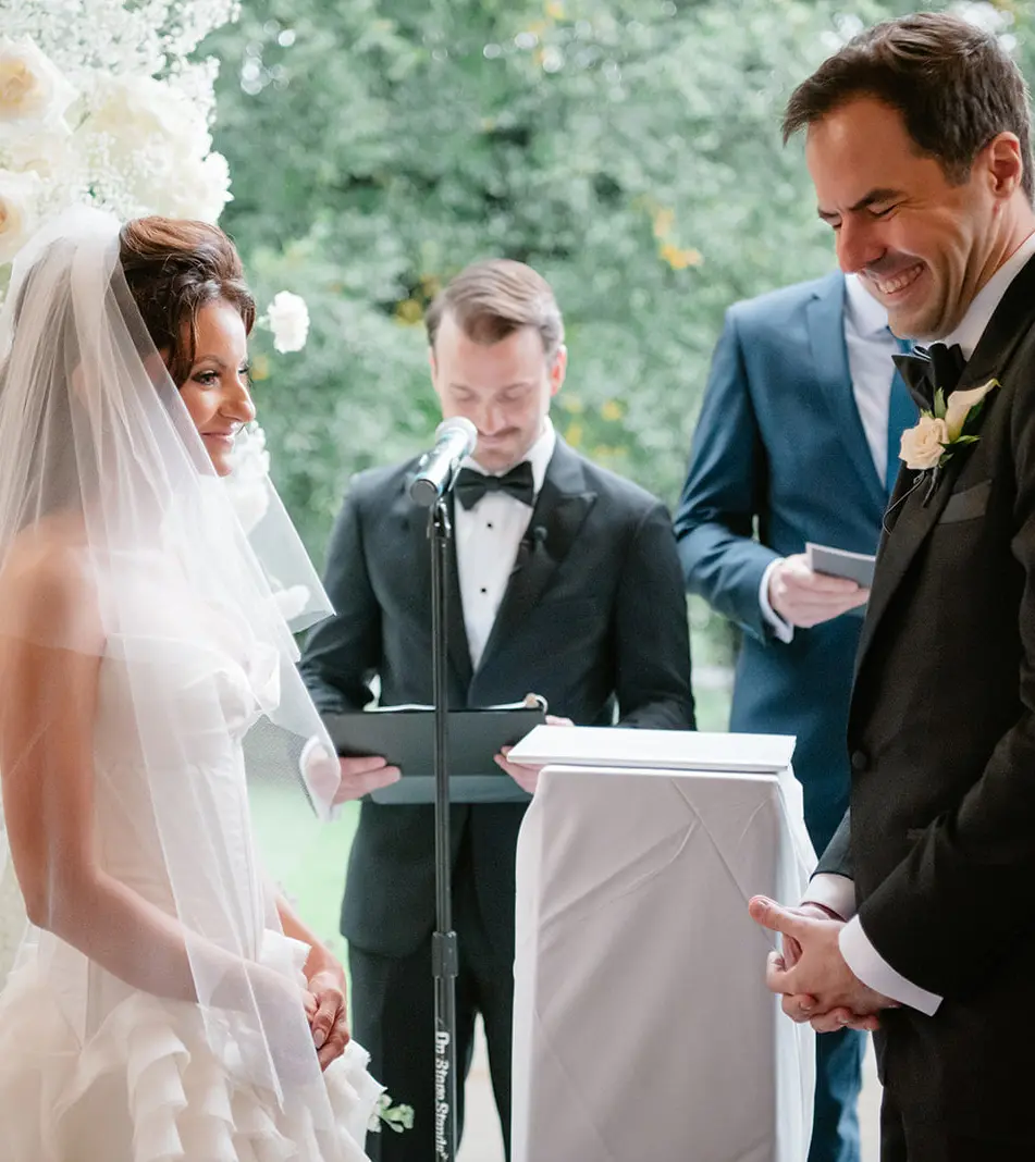 On the porch, between walls of white flowers, the bride and groom face each other as the officiant reads.