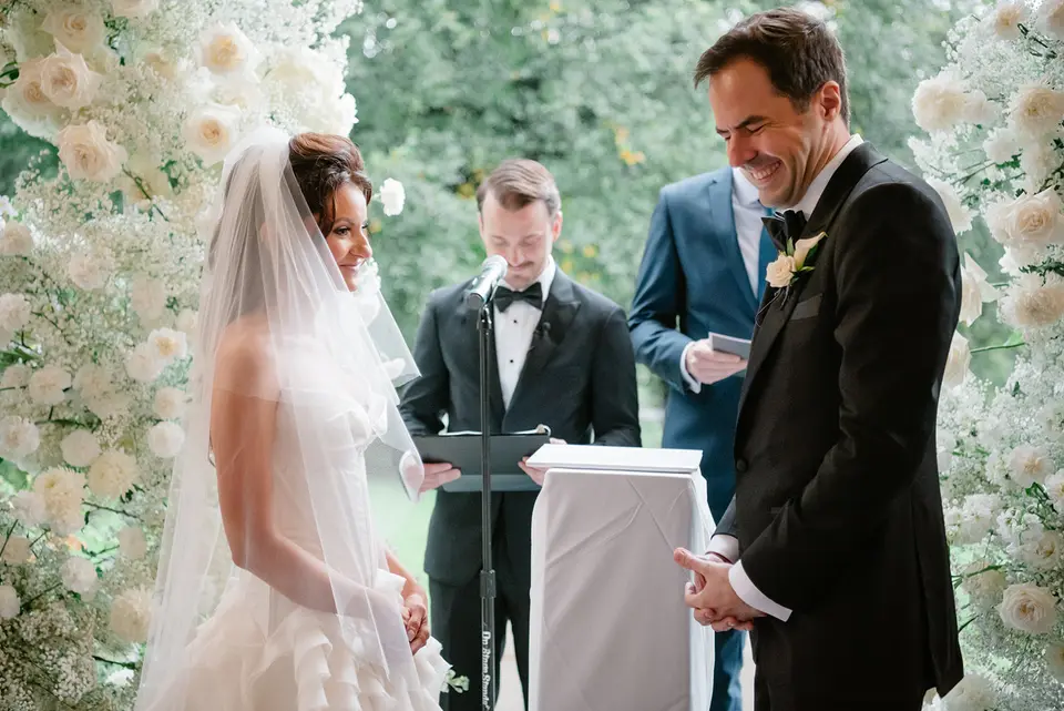 On the porch, between walls of white flowers, the bride and groom face each other as the officiant reads.
