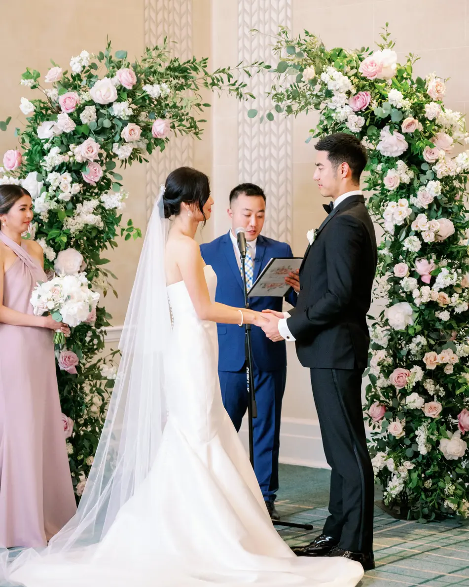 Beneath the arch of flowers, the couple hold hands before their officiant, a bridesmaid in blush beside them.