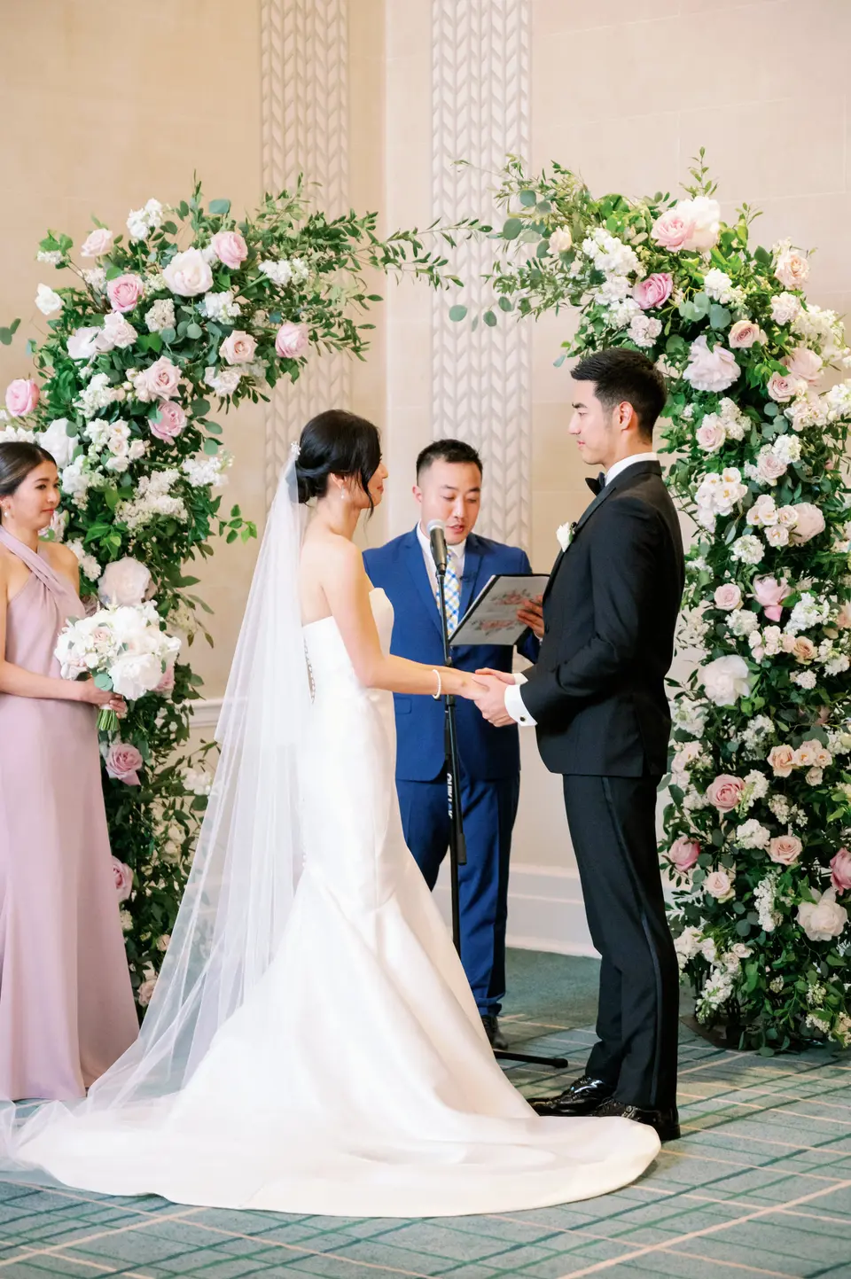 Beneath the arch of flowers, the couple hold hands before their officiant, a bridesmaid in blush beside them.