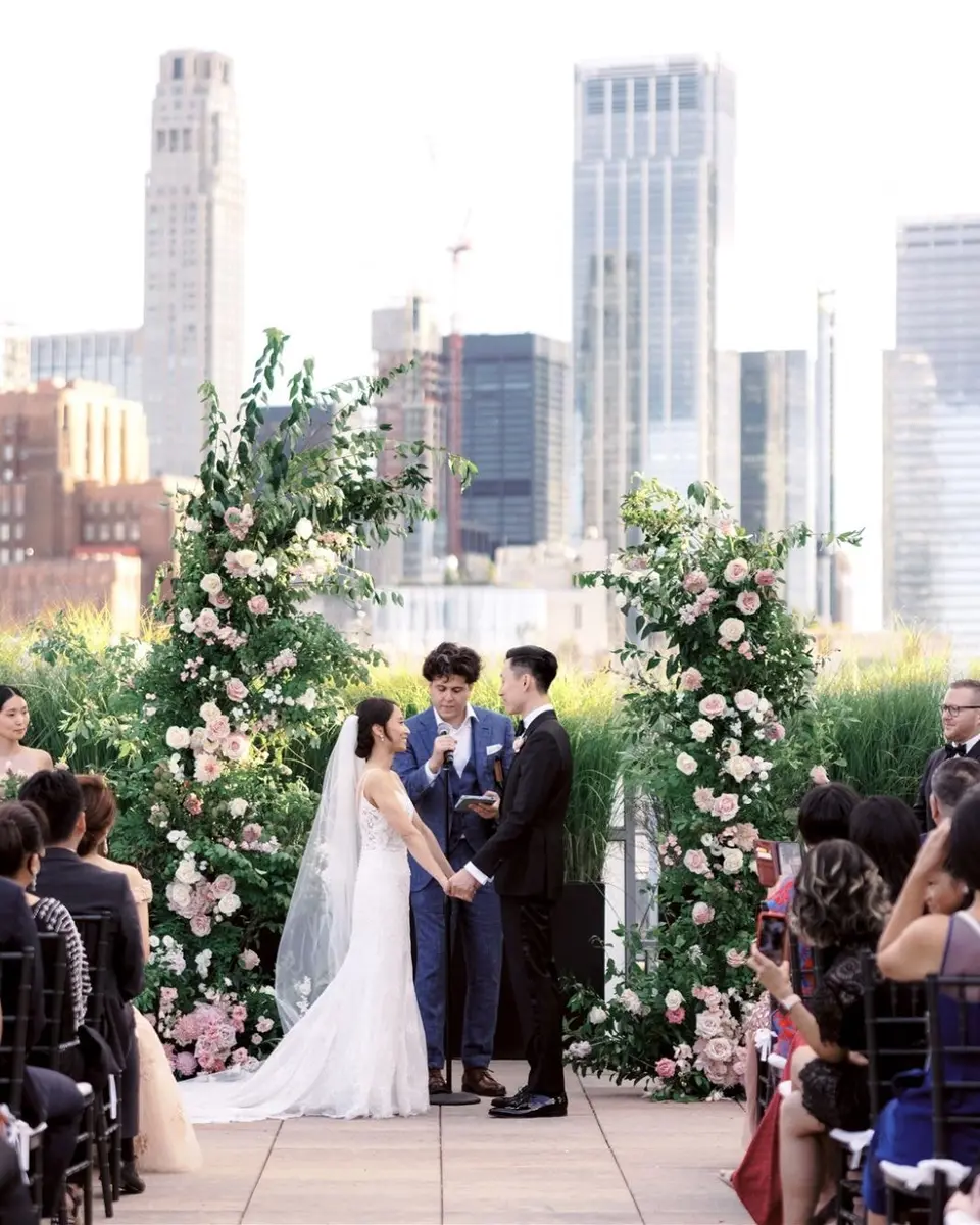 The bride and groom hold hands as they exchange vows between two tall arches of greenery and blush roses on the Tribeca Rooftop, the officiant between them and the Lower Manhattan skyline behind.