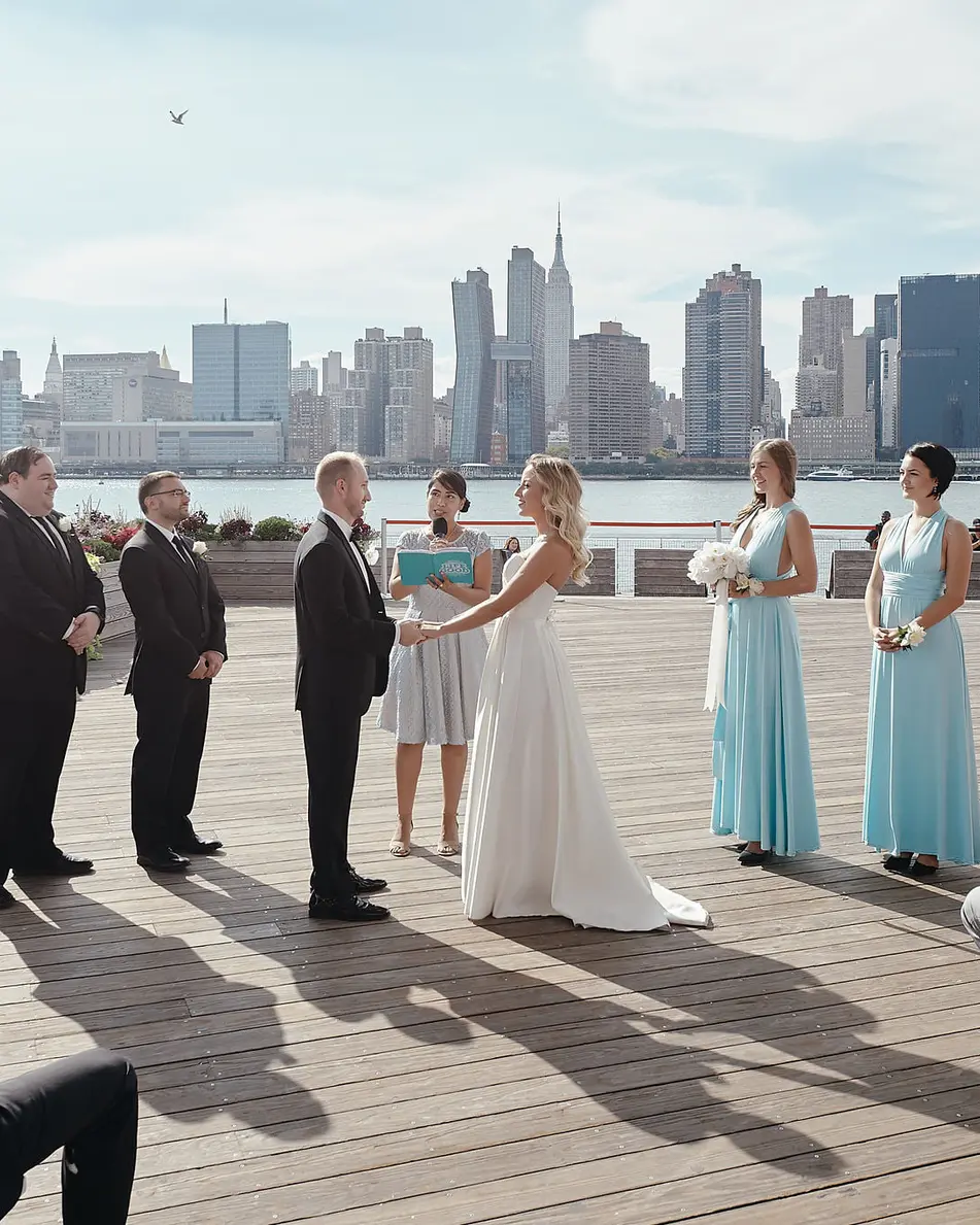 The couple stand before their officiant on the deck, bridesmaids in aqua beside them and the skyline across the water.