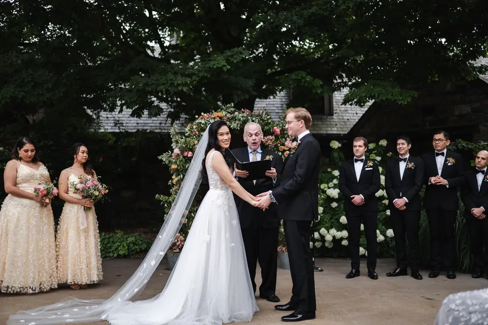 In the stone courtyard beneath tall trees, the couple hold hands before their officiant, bridesmaids in champagne on one side and groomsmen on the other.
