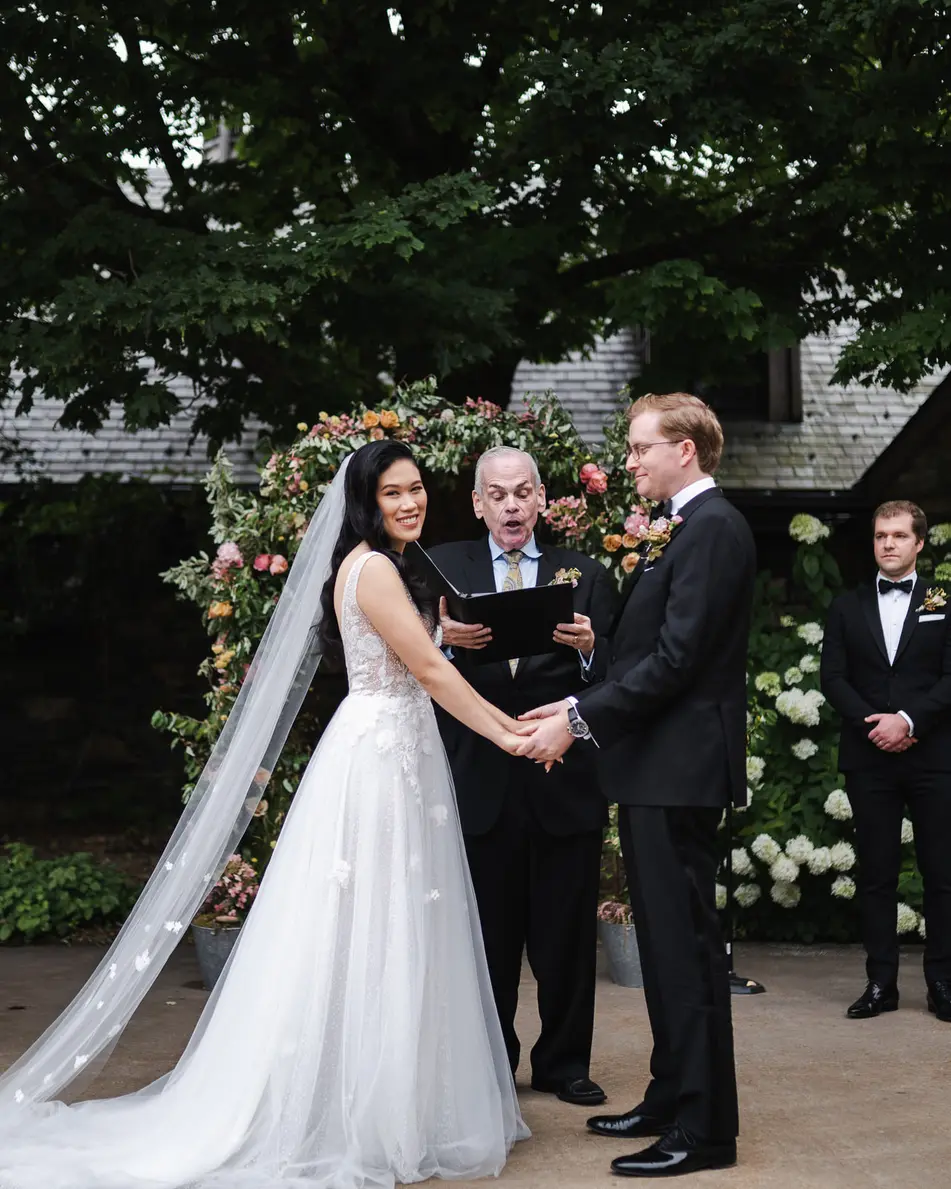 In the stone courtyard beneath tall trees, the couple hold hands before their officiant, bridesmaids in champagne on one side and groomsmen on the other.