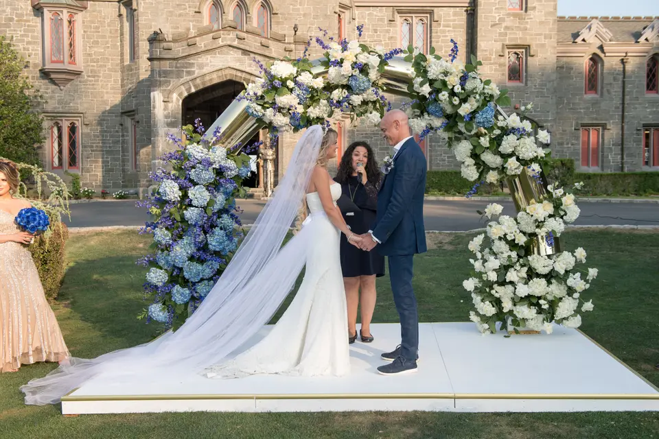 The couple hold hands beneath the arch of blue and white flowers before the castle's stone doorway.