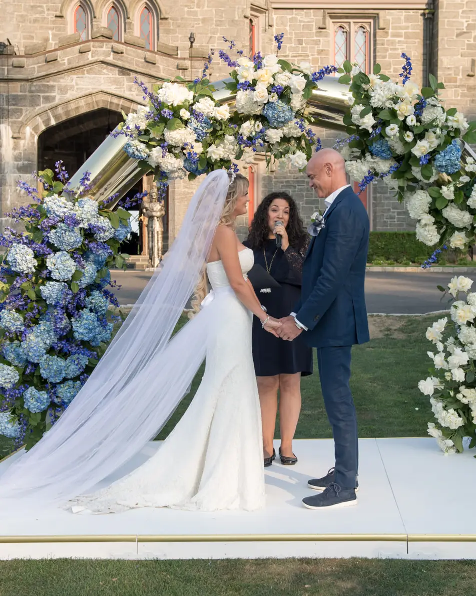 The couple hold hands beneath the arch of blue and white flowers before the castle's stone doorway.