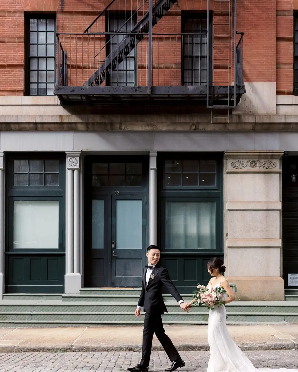 The couple walk hand in hand along a Tribeca street in front of a red brick warehouse with green iron doors.