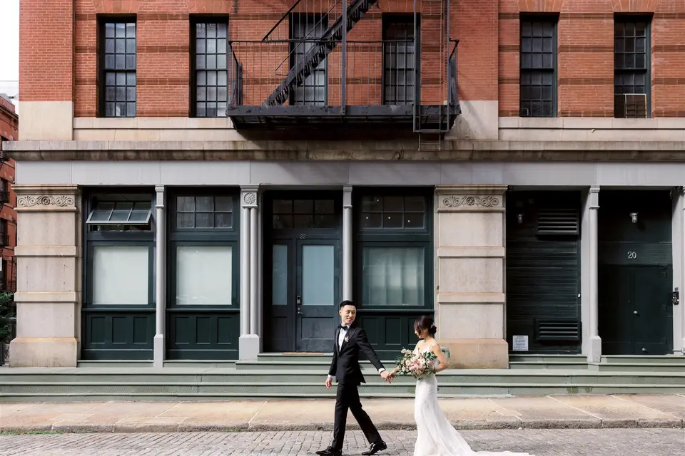 The couple walk hand in hand along a Tribeca street in front of a red brick warehouse with green iron doors.