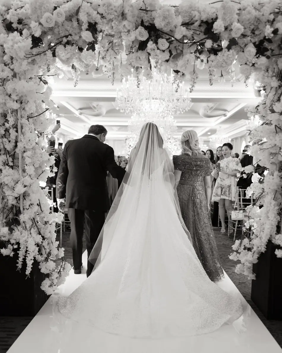 From behind, the bride walks toward the ceremony beneath an arch of white flowers and crystal. Black and white.