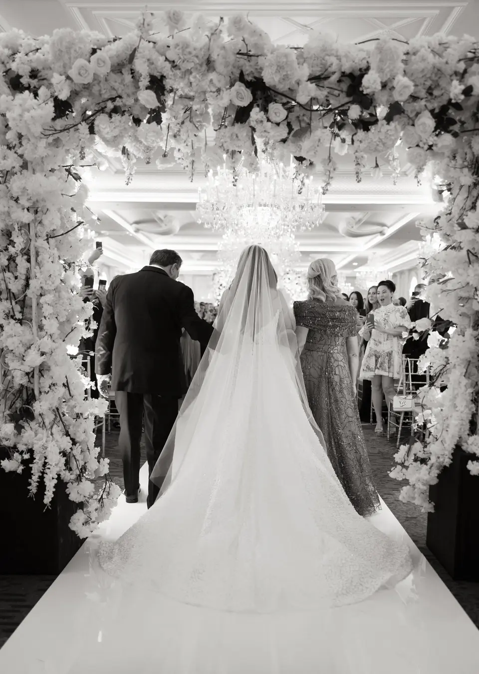 From behind, the bride walks toward the ceremony beneath an arch of white flowers and crystal. Black and white.