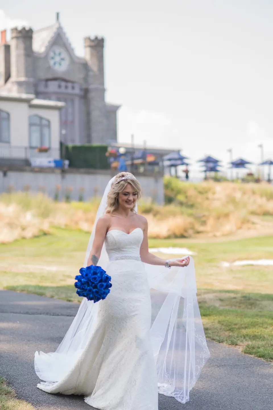 The bride walks across the lawn with a bouquet of blue roses, the stone castle behind her.