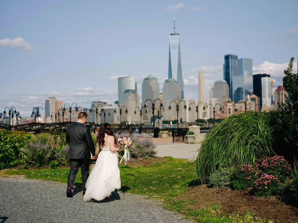 The couple walk away along a garden path at Liberty State Park, hand in hand, the towers of Lower Manhattan across the river.