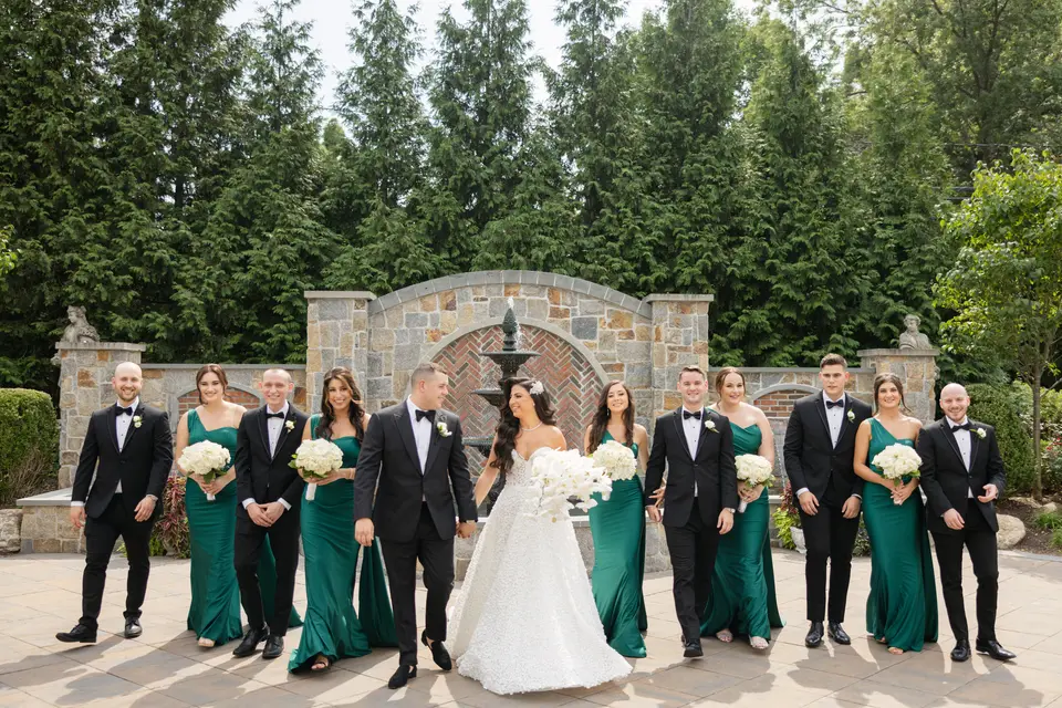 The wedding party walk toward the camera across a stone courtyard, a fountain and tall evergreens behind them.