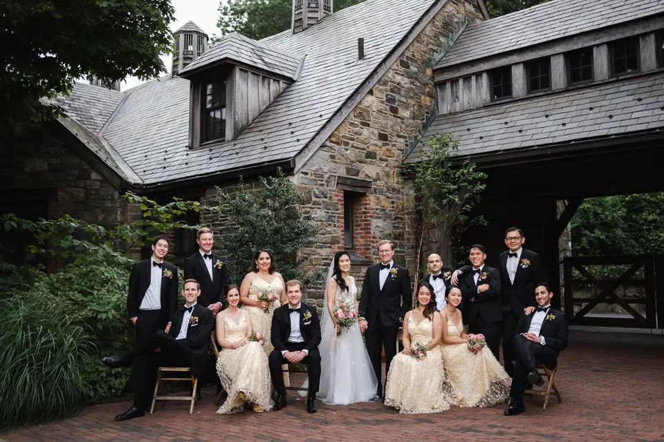 The wedding party sit and stand together in a brick courtyard before the stone barns, bridesmaids in champagne gowns.