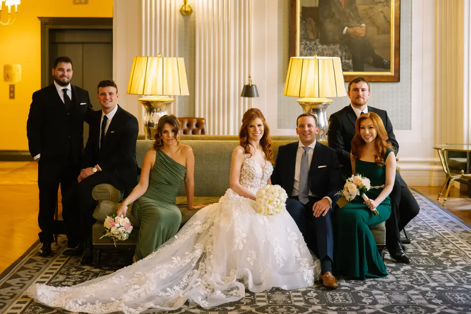 The wedding party gathered on a green sofa in a lamp-lit lounge, the bride at the center in a lace gown.