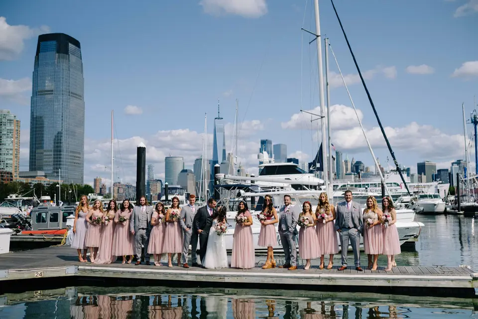 The wedding party lines the dock of a marina, sailboats beside them and the Jersey City and Manhattan towers beyond.
