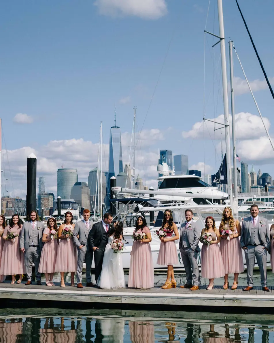 The wedding party lines the dock of a marina, sailboats beside them and the Jersey City and Manhattan towers beyond.