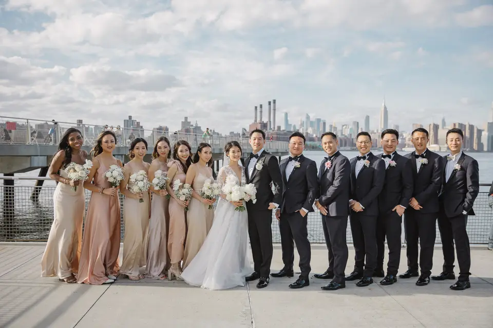 The whole wedding party stand in a line on the waterfront, the skyline and the Empire State Building behind them.