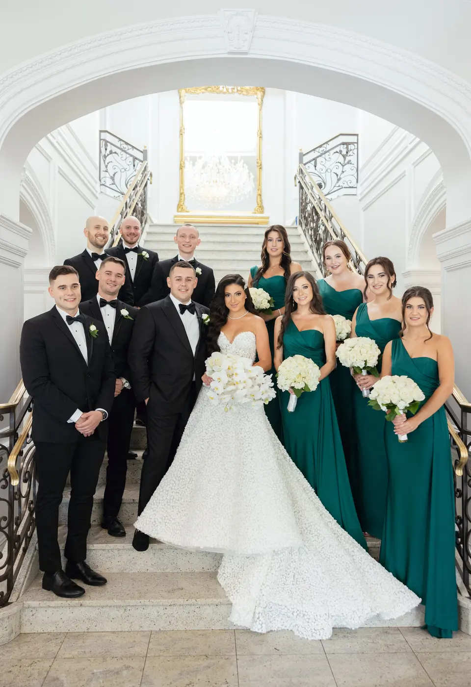 The wedding party on the grand staircase, bridesmaids in emerald and groomsmen in black tie around the couple.