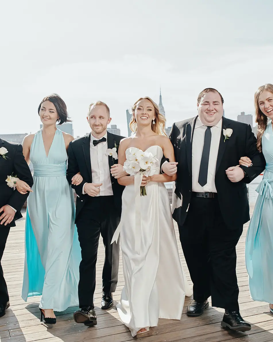 The wedding party walks arm in arm along the waterfront, bridesmaids in aqua and groomsmen in black tie.