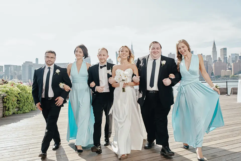 The wedding party walks arm in arm along the waterfront, bridesmaids in aqua and groomsmen in black tie.