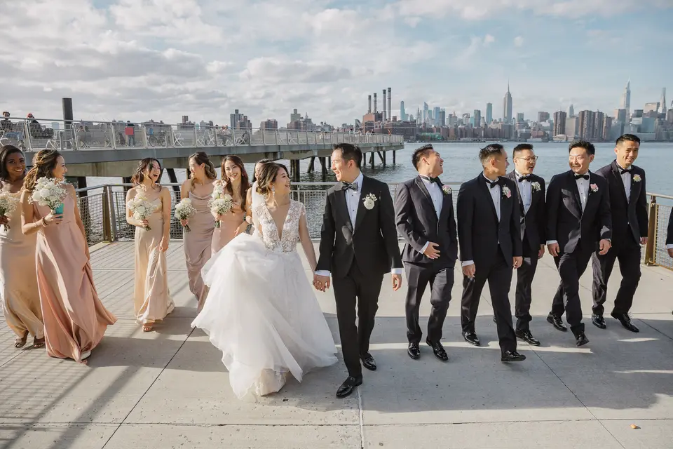 The wedding party walk along the East River waterfront, bridesmaids in champagne satin, the Manhattan skyline behind them.