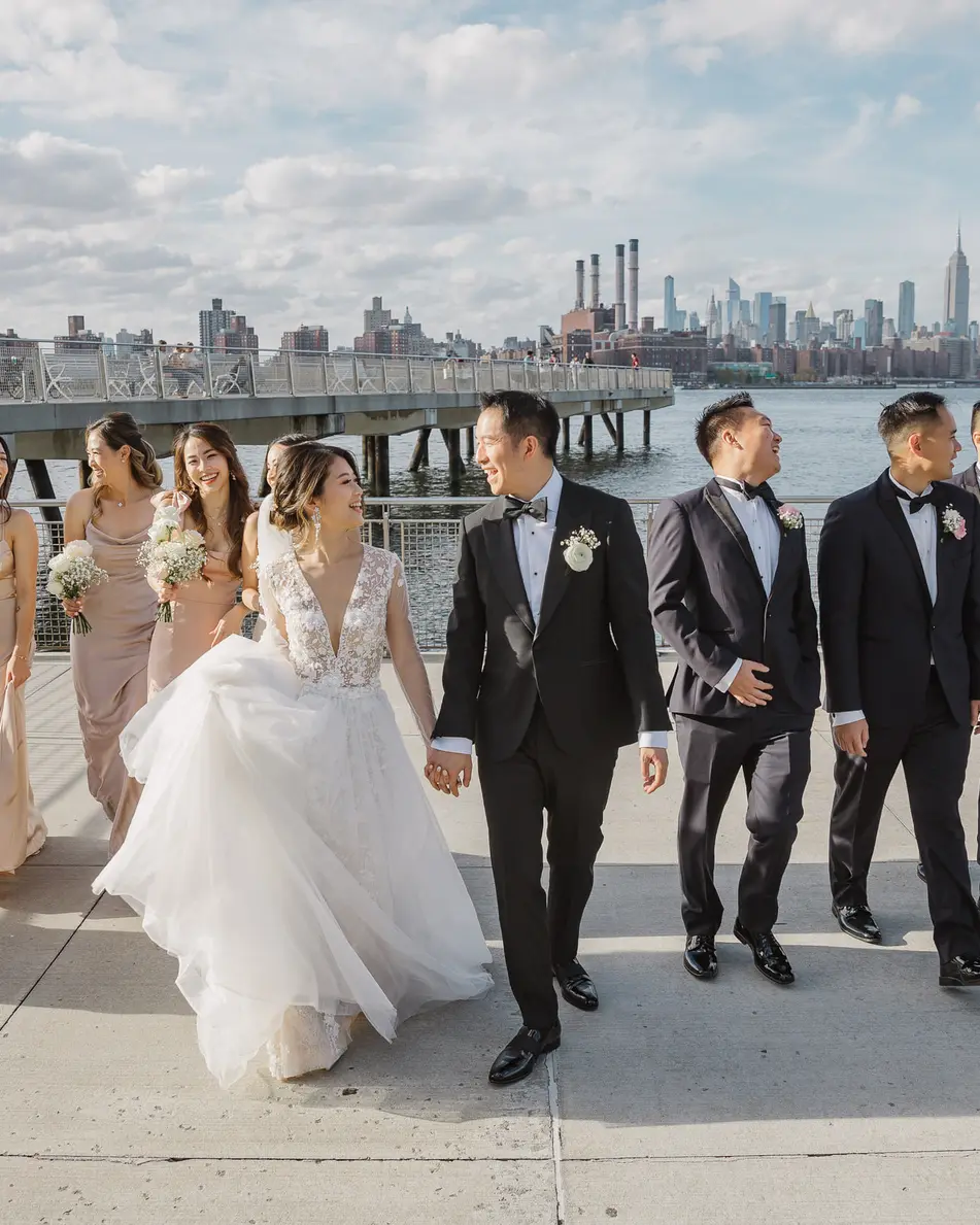 The wedding party walk along the East River waterfront, bridesmaids in champagne satin, the Manhattan skyline behind them.
