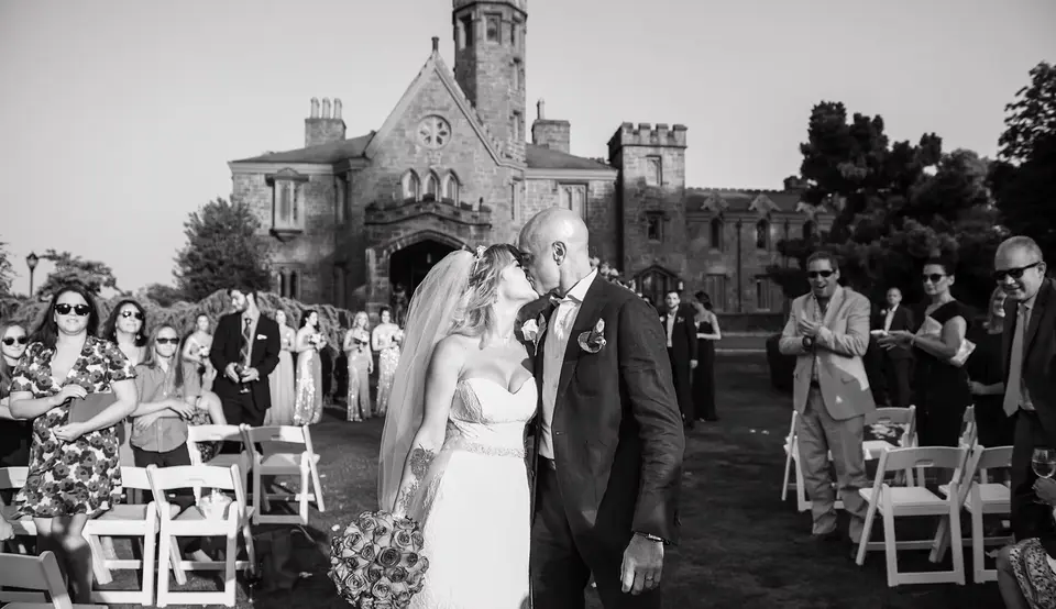 Guests stand and applaud as the couple walk back up the aisle on the lawn at Whitby Castle, the stone castle behind them. Black and white.