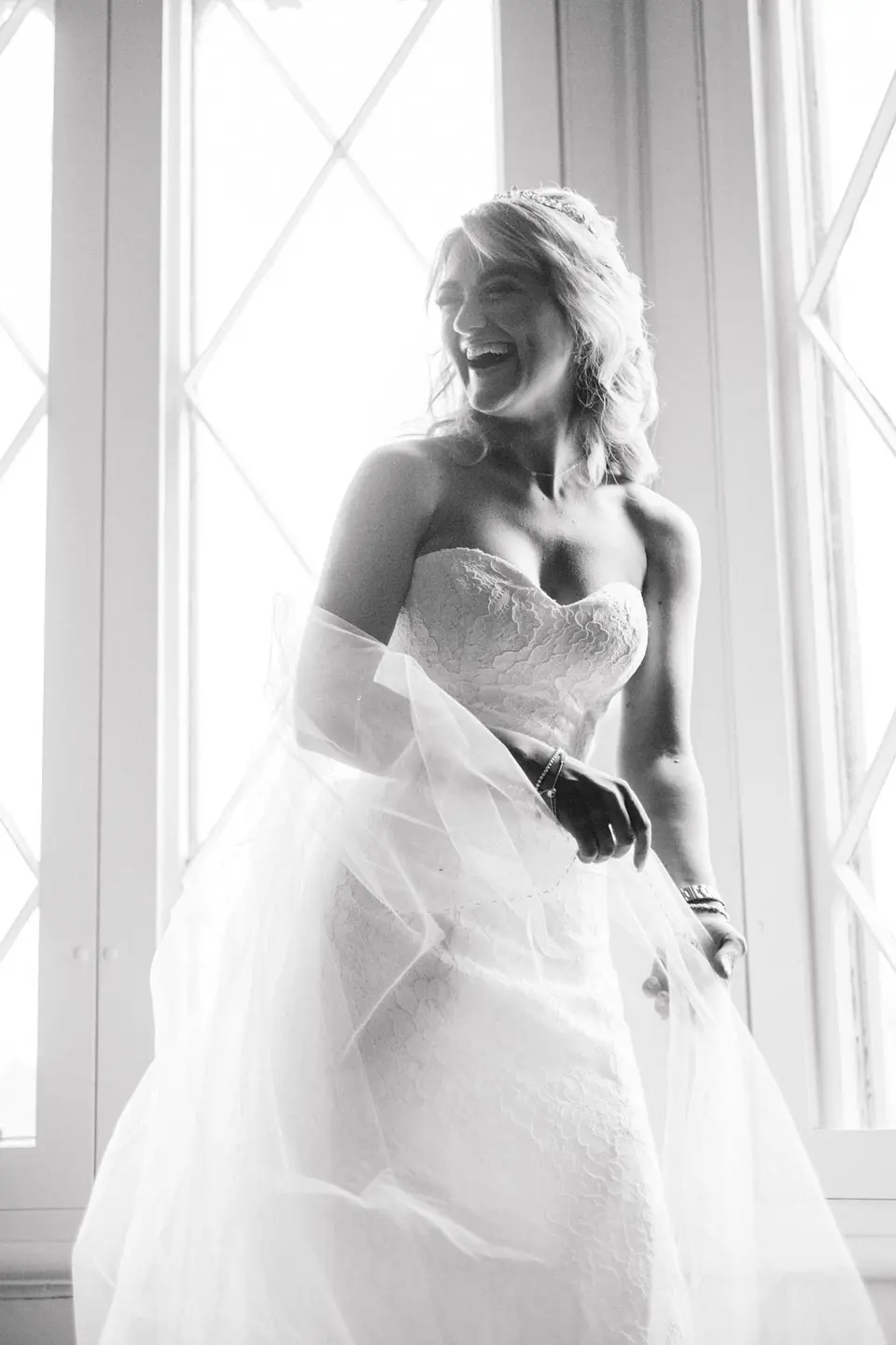 The bride laughs by a tall window, lifting her skirt. Black and white.