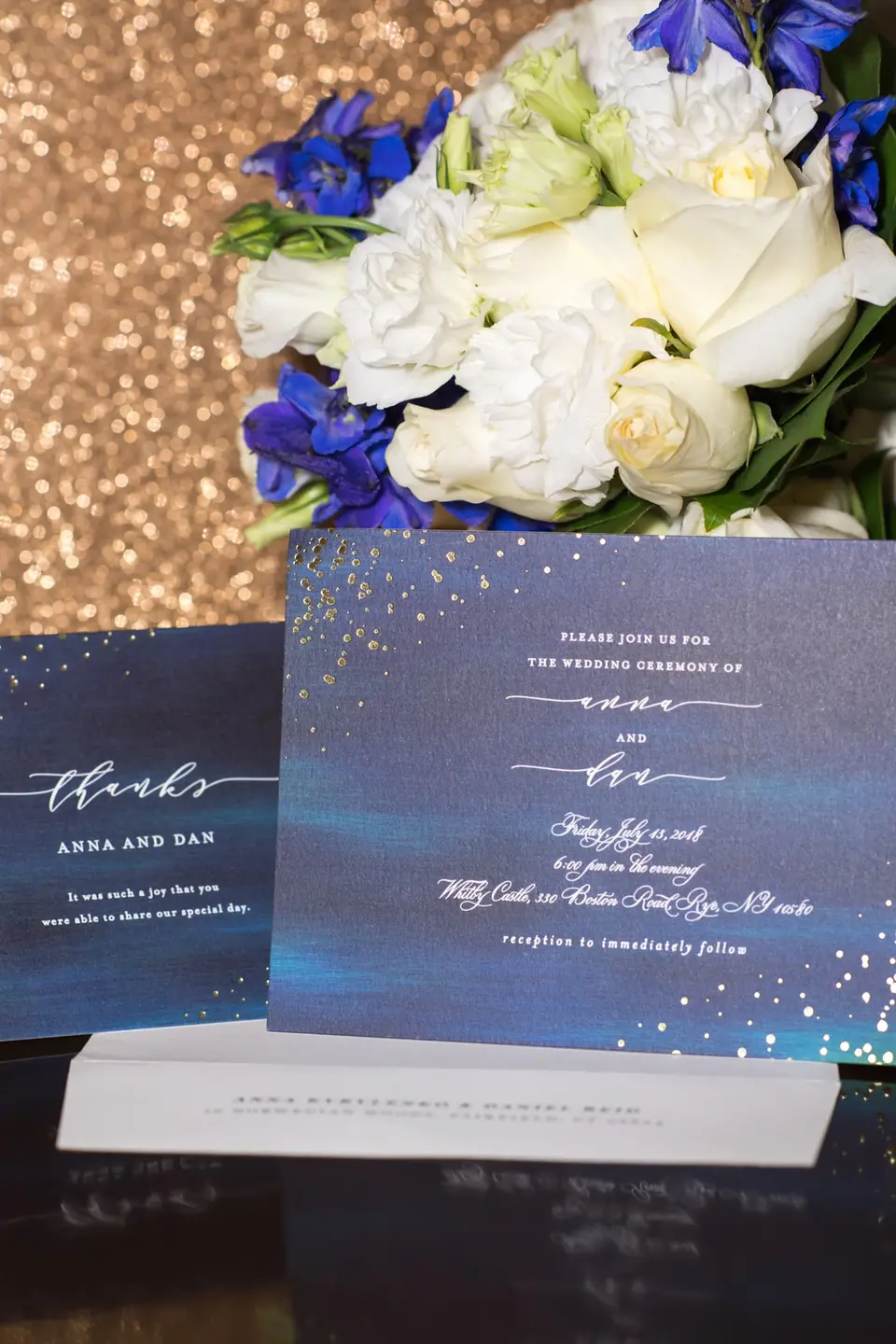 The invitation and thank-you card in deep blue with gold flecks, beneath white roses and blue delphinium.