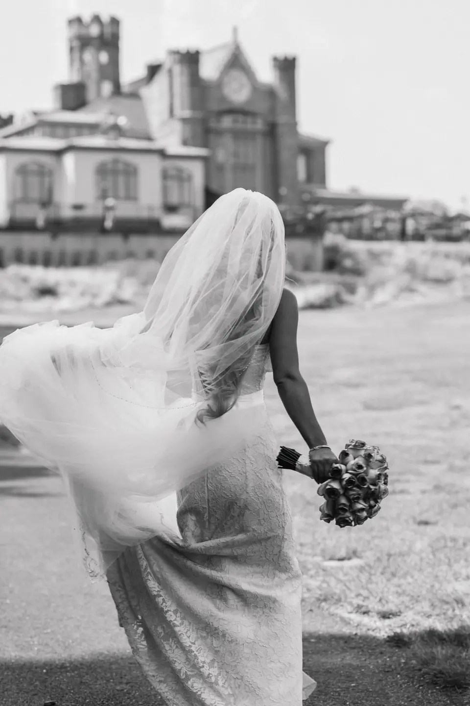 The bride walks across the lawn toward the castle, her veil blowing. Black and white.