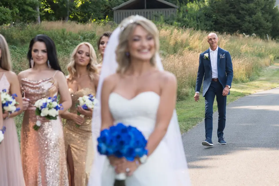 The groom walks up the path toward the bride for the first look, her bridesmaids in gold beside her.